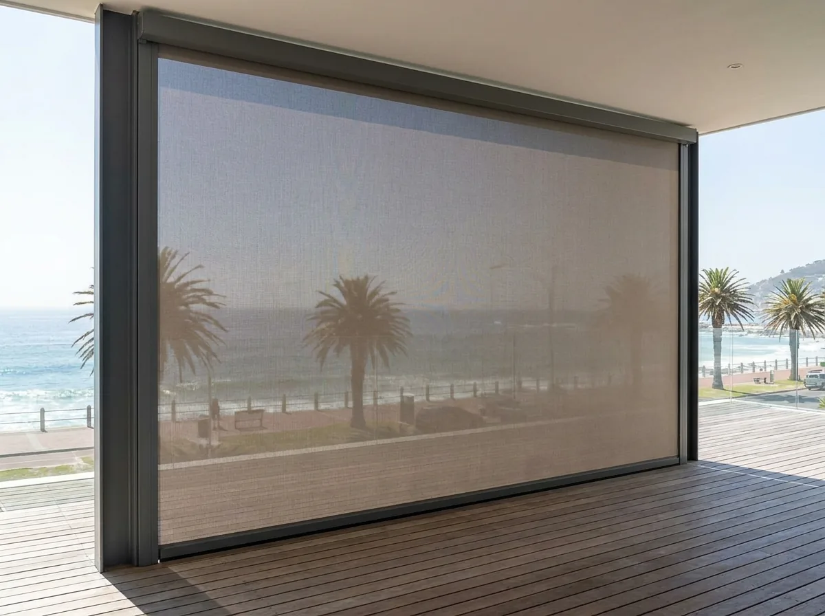 A single flat exterior zip screen lowered taut in fixed side channels over the open side of a Camps Bay deck, the mesh filtering the view to the Atlantic Ocean and palm-lined beachfront promenade